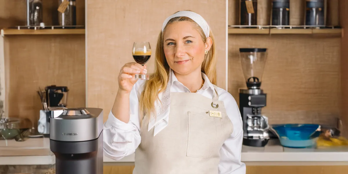 Female Nespresso Employee Smiles While Holding a Cup of Coffee Next to a Coffee Maker in a Kitchen Setting.
