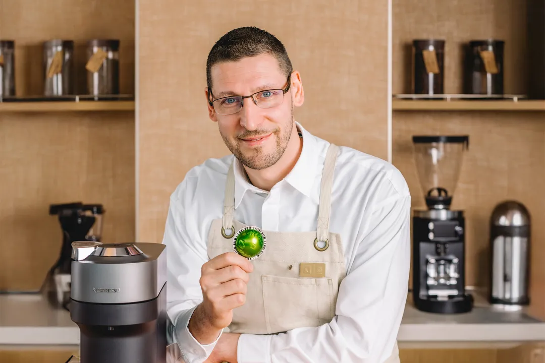 A barista in an apron holding a green coffee pod next to a coffee machine.