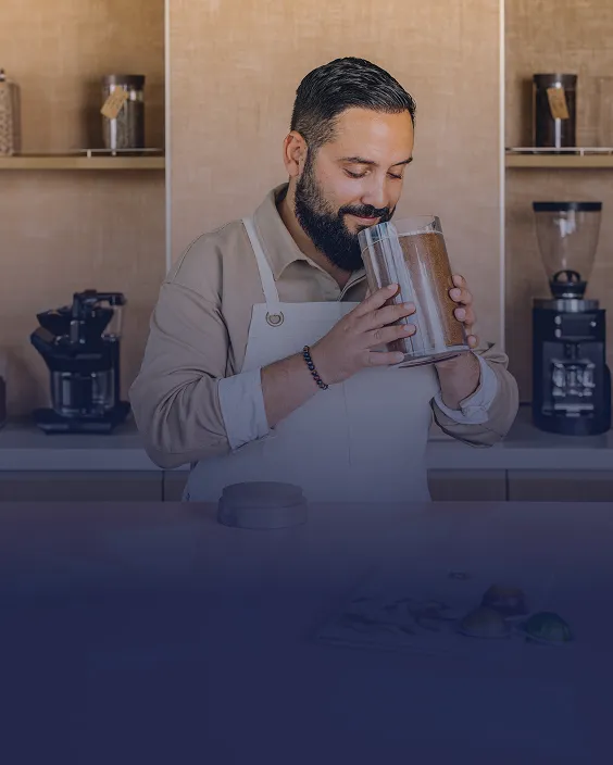 A man in an apron holds a container filled with coffee granules in a kitchen setting.